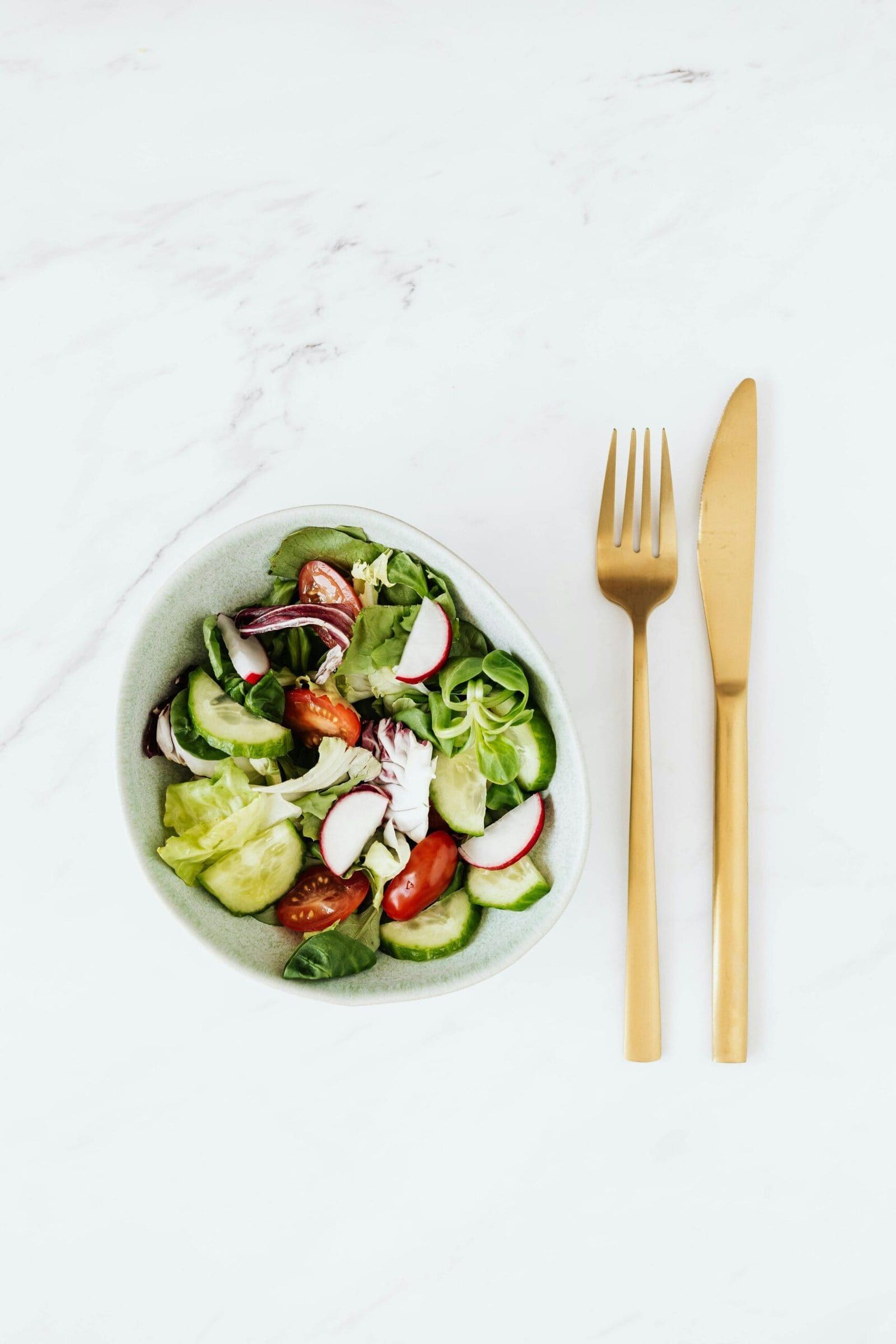 Bowl of fresh vegetable salad with golden cutlery on a marble surface, healthy meal concept.